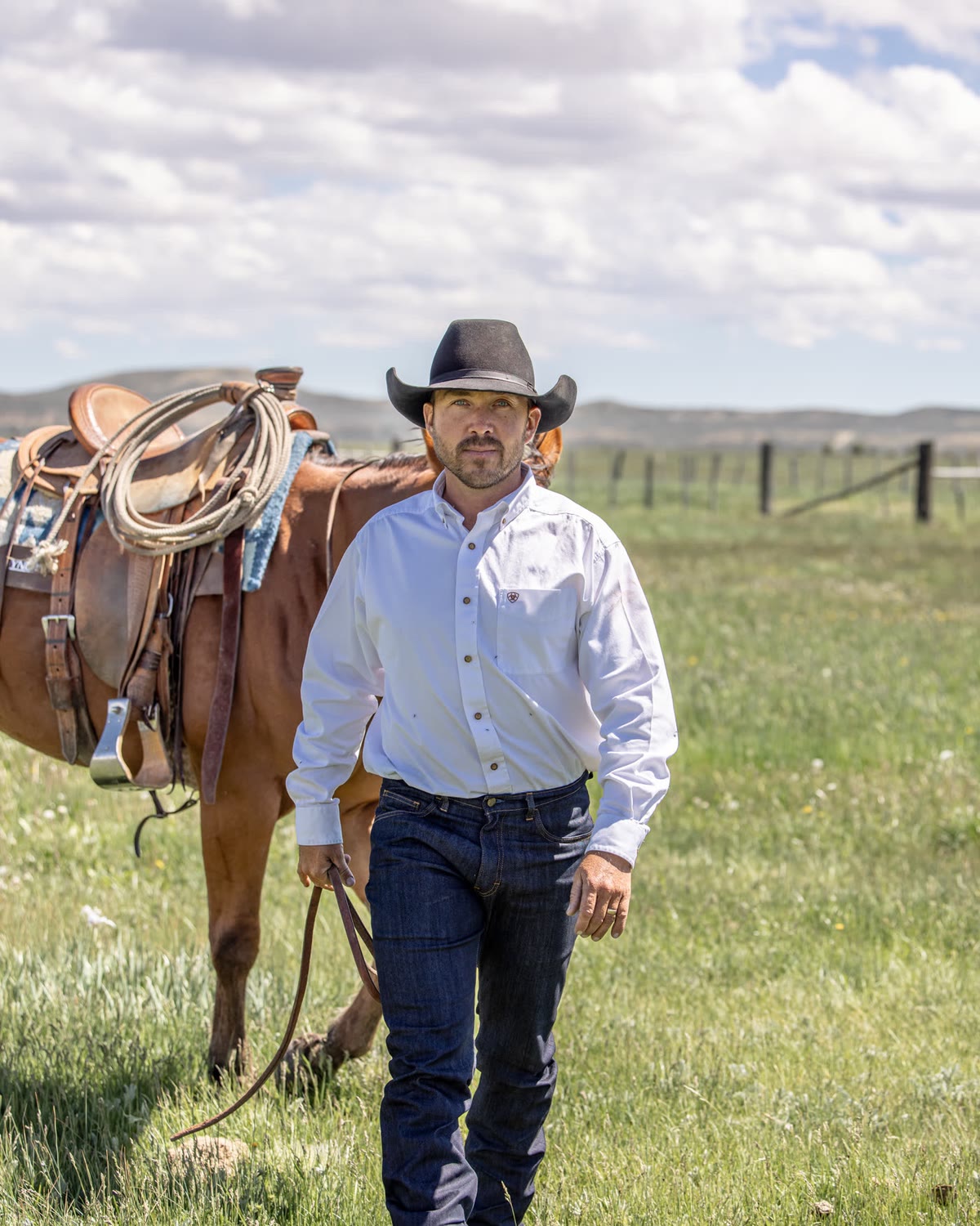 Man in a white shirt and dark jeans walking beside a saddled horse