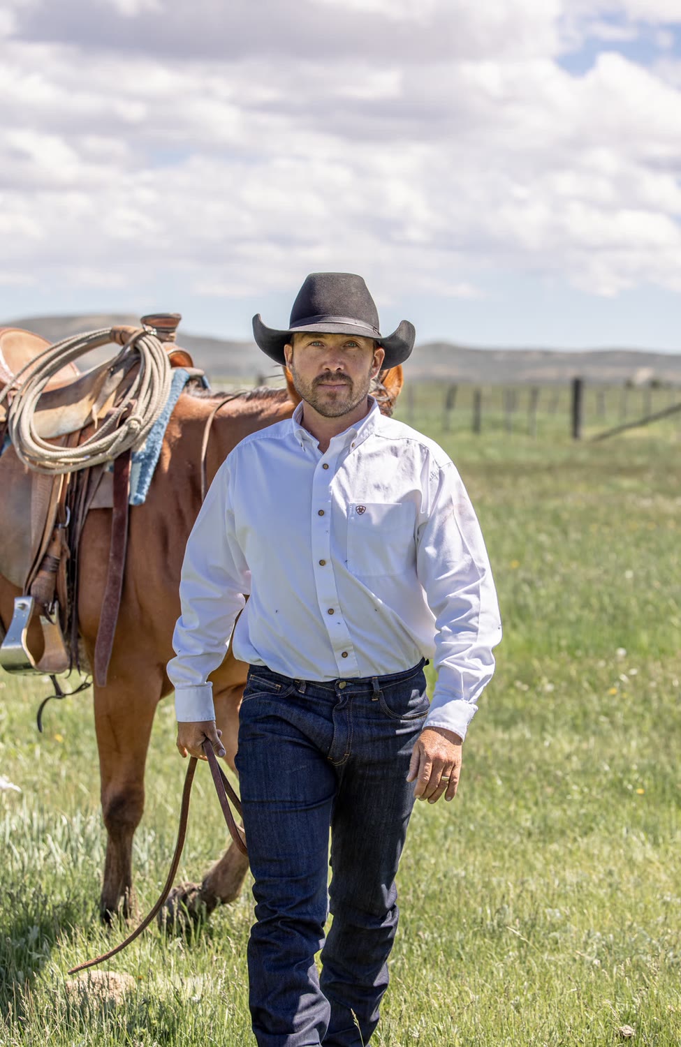 Man in a white shirt and dark jeans walking beside a saddled horse