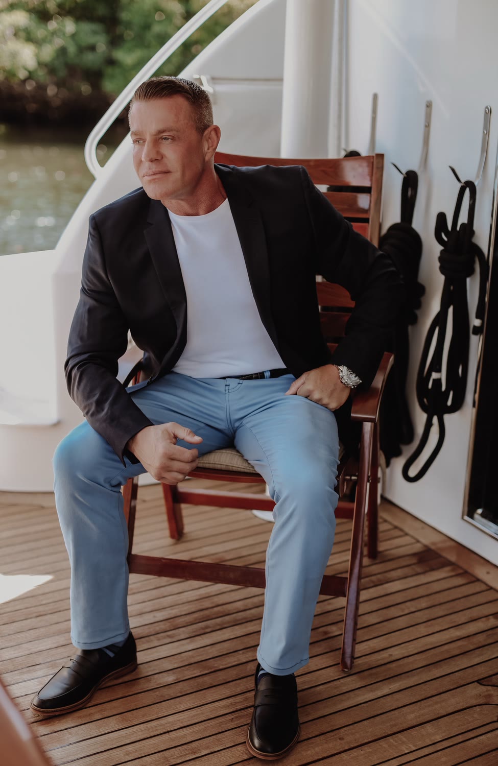 Man in a navy blazer, white tee and light-wash jeans seated on the deck of a boat