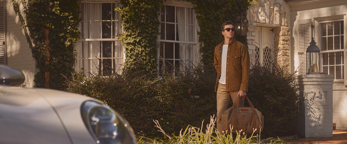 A man in khaki pants carrying a waxed canvas duffle outside an ivy-covered house