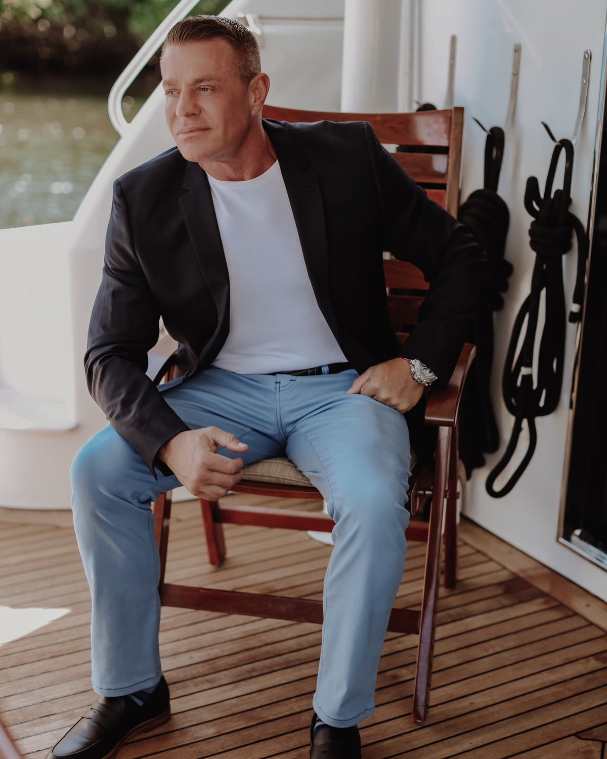 Man in a navy blazer, white tee and light-wash jeans seated on the deck of a boat