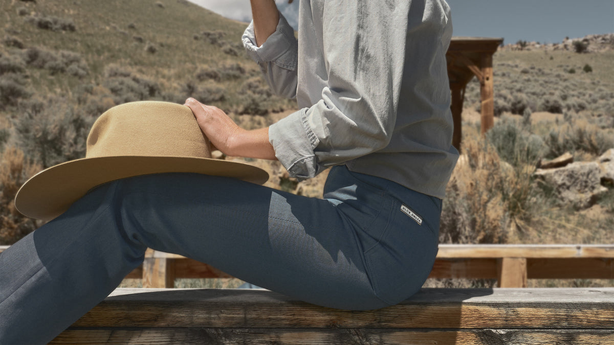 Woman in a denim shirt and blue flare jeans seated on a rail, her hat resting on her knee