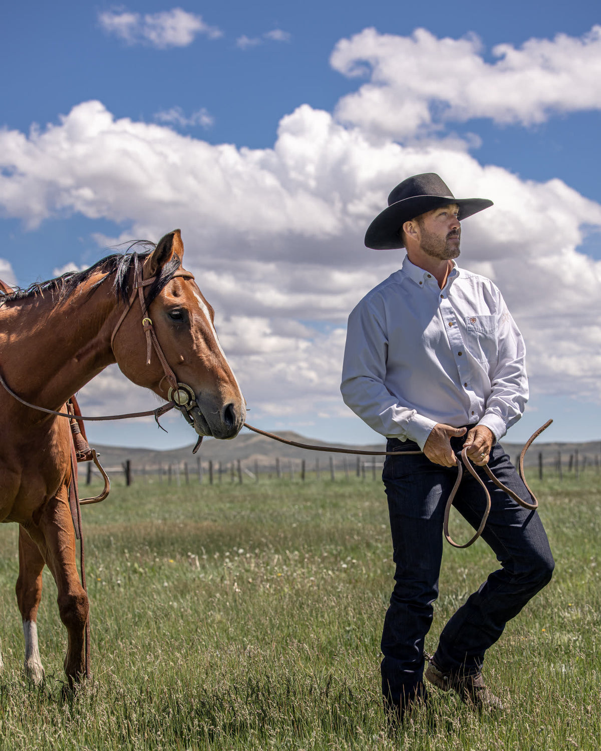 Man in dark raw denim jeans and a cowboy hat standing with a horse on open range
