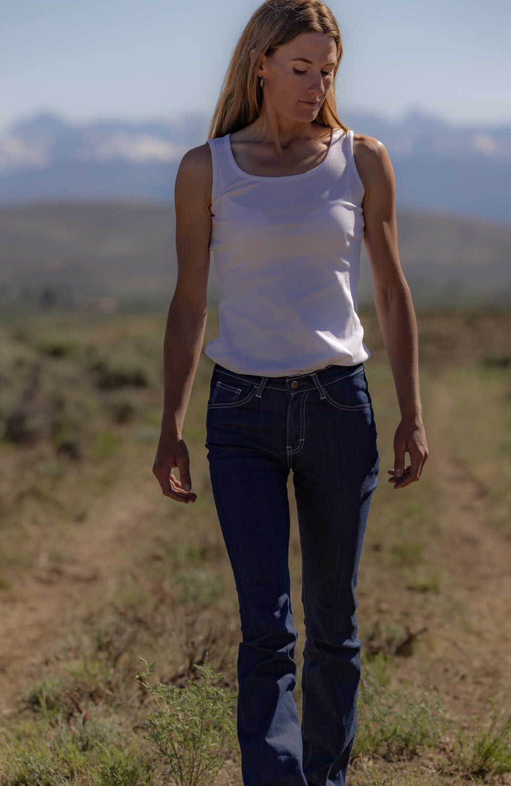 Woman in raw denim jeans and a white tank top walking a dirt road through open grassland