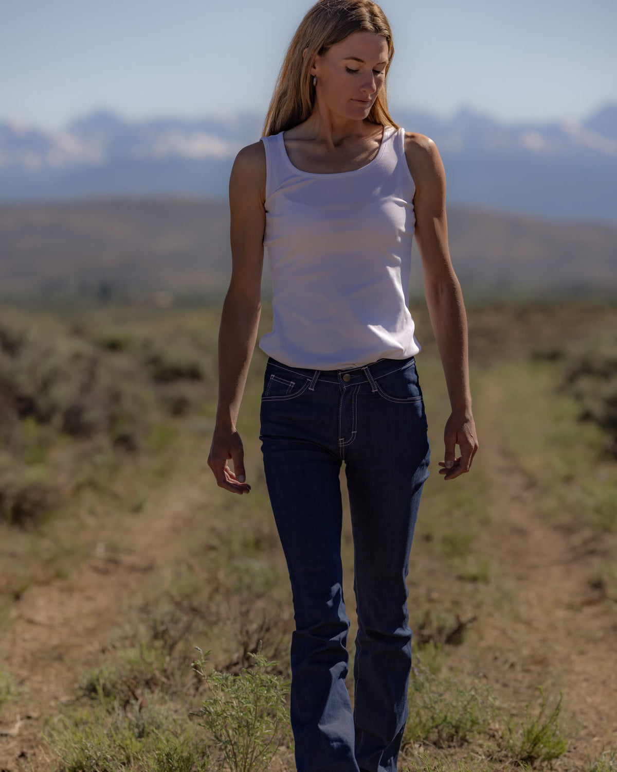 Woman in raw denim jeans and a white tank top walking a dirt road through open grassland