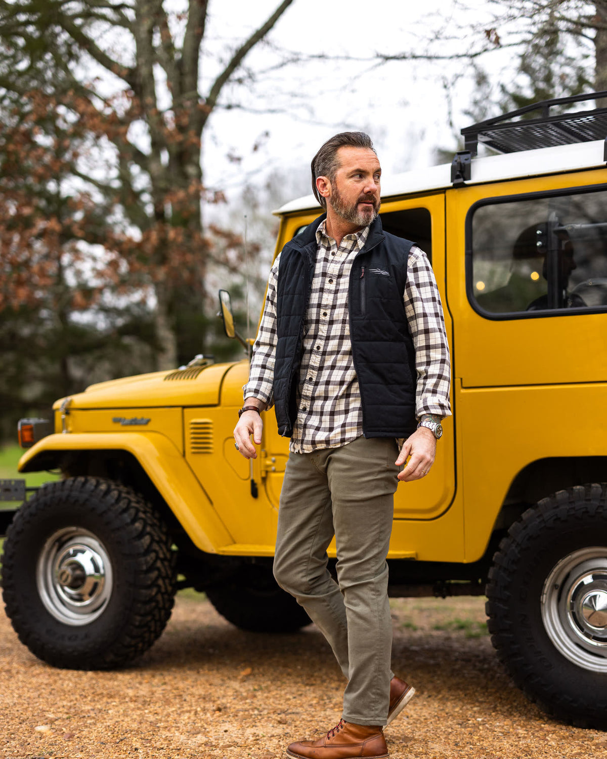 Man in Banana Olive cotton chinos, a plaid shirt and a quilted vest beside a yellow truck