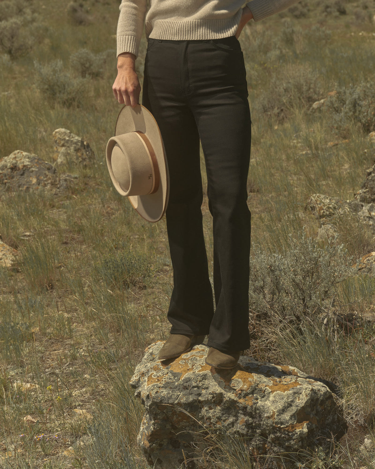 Woman in black flare jeans standing on a rock in the sagebrush, hat in hand