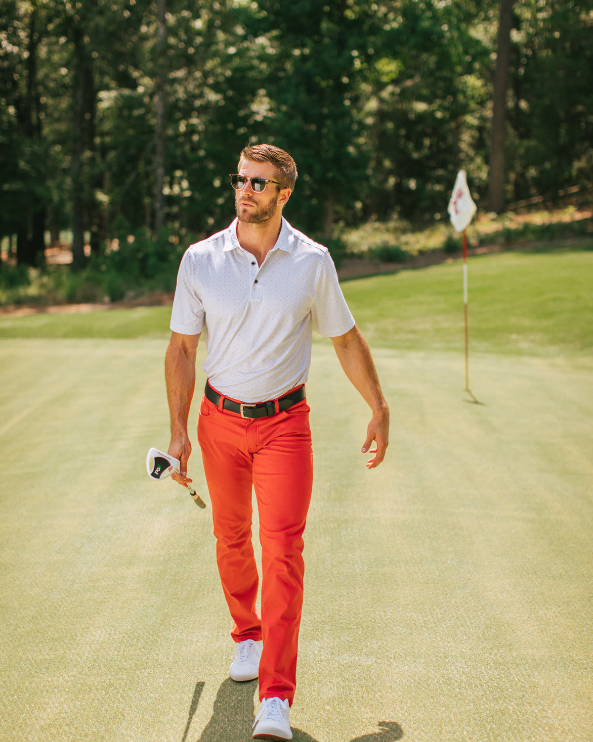 Man in red performance pants and a white polo walking a golf green with a club