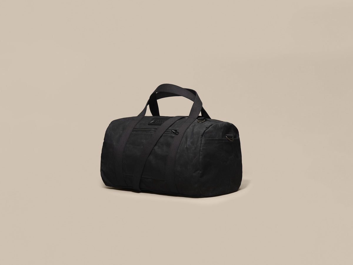 Waxed Canvas Duffle in black waxed canvas with black webbing and a black leather patch, angled view