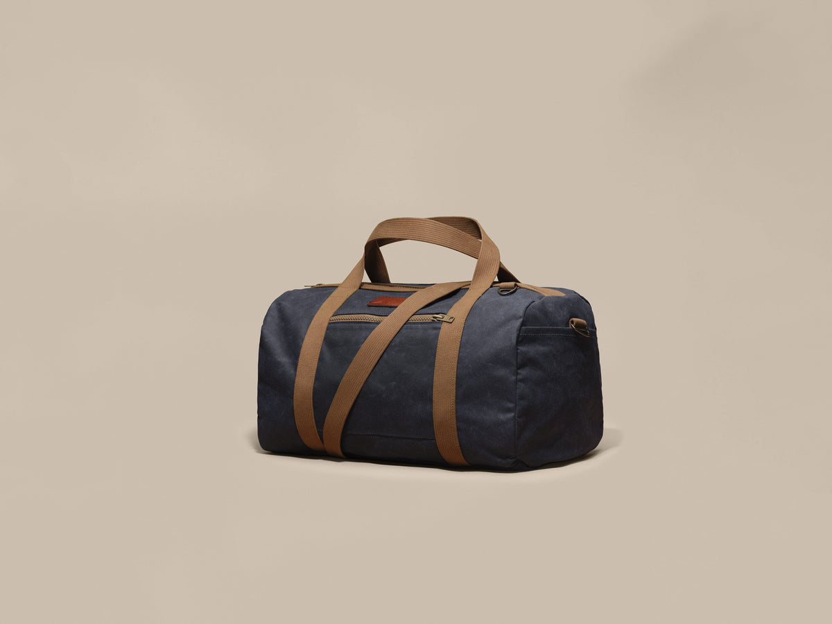 Waxed Canvas Duffle in navy waxed canvas with tan webbing and a Rich Brown leather patch, angled view