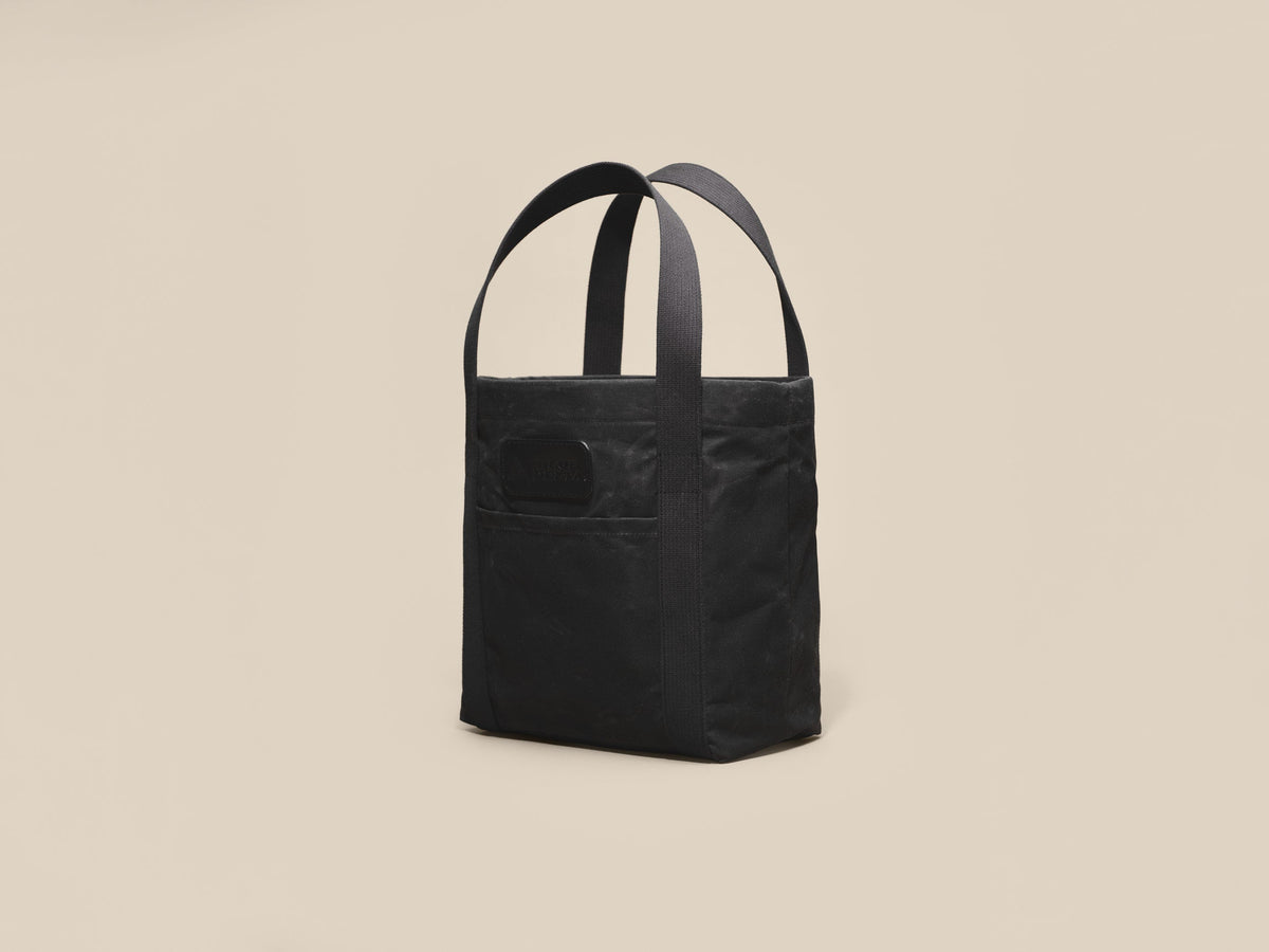 Waxed Canvas Tote in black waxed canvas with black webbing and a black leather patch, angled view