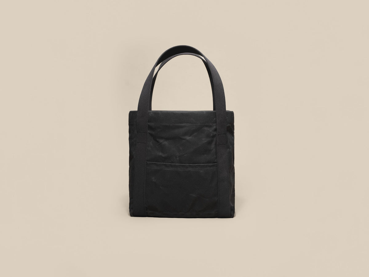 Waxed Canvas Tote in black waxed canvas with black webbing and a black leather patch, slip pocket side